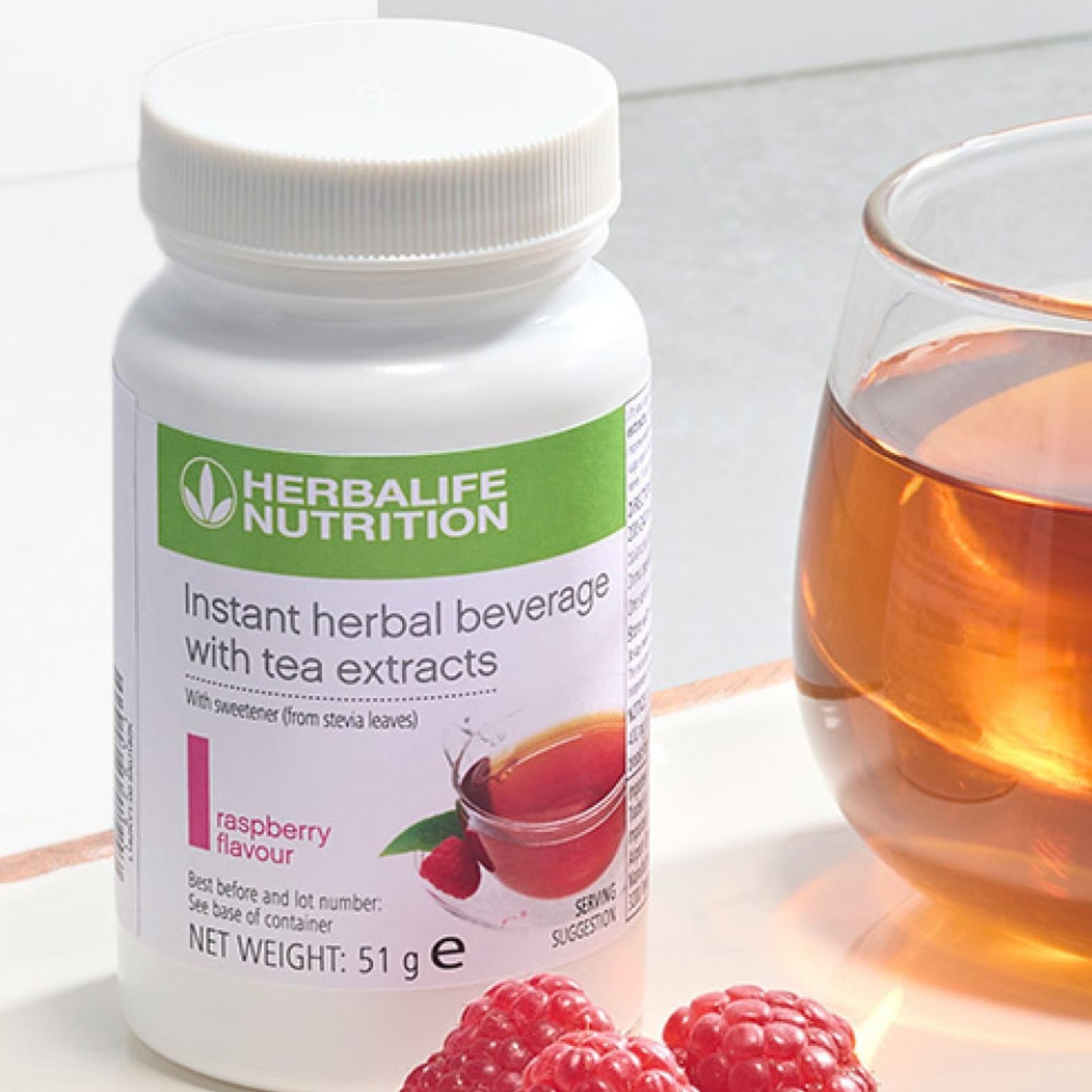 Herbalife Instant Herbal Beverage raspberry 51 g tub beside a glass of prepared tea and fresh raspberries