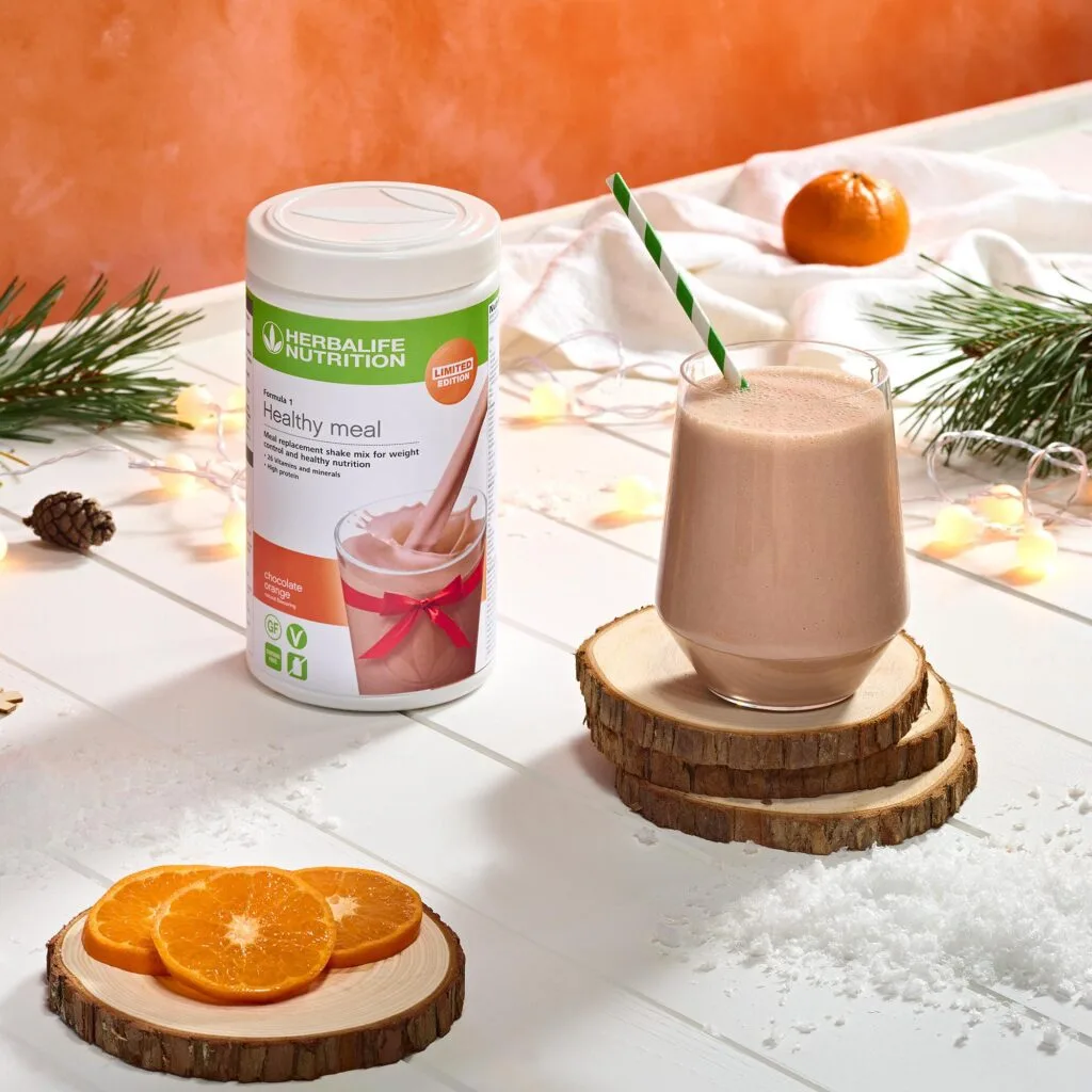 Herbalife Formula 1 Healthy Meal chocolate orange shake mix with a prepared smoothie glass on wooden coasters and fresh orange slices.