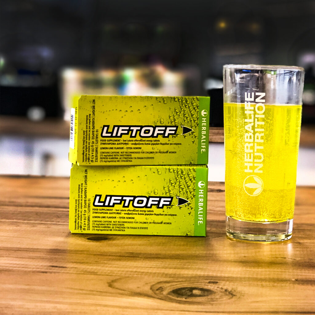 Two stacked green boxes of Herbalife Liftoff Lemon-Lime energy tablets next to a tall glass of sparkling yellow effervescent drink on a wooden table.