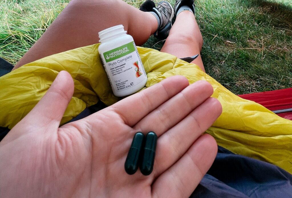 Hand holding two dark green capsules in front of an open bottle of Herbalife Phyto Complete food supplement in an outdoor camping setting.