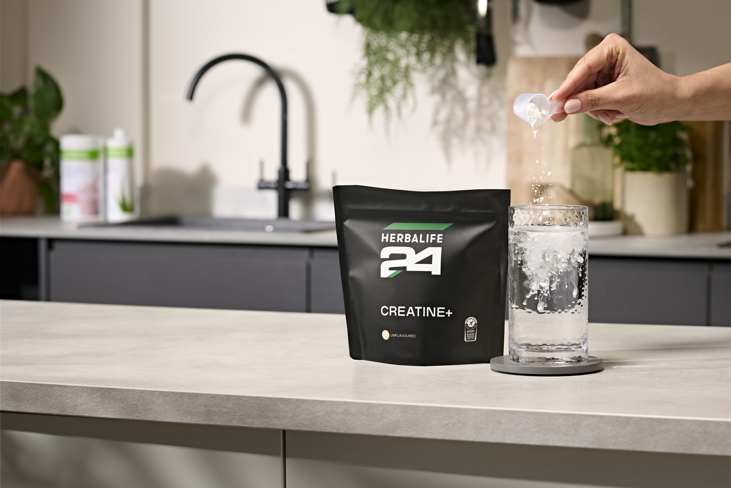 A hand pouring a scoop of white Herbalife24 Creatine Plus powder into a tall glass of effervescent water next to the black product pouch on a modern kitchen counter.