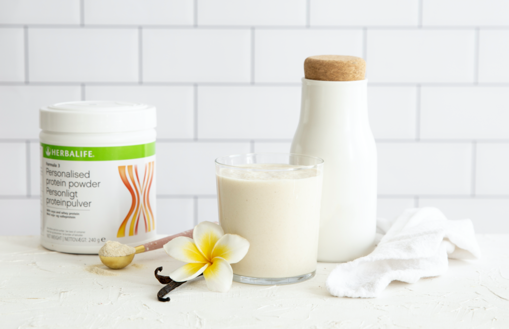 Herbalife Formula 3 Personalised Protein Powder canister next to a freshly blended vanilla protein shake glass with a plumeria flower and a white milk carafe on a textured white surface.