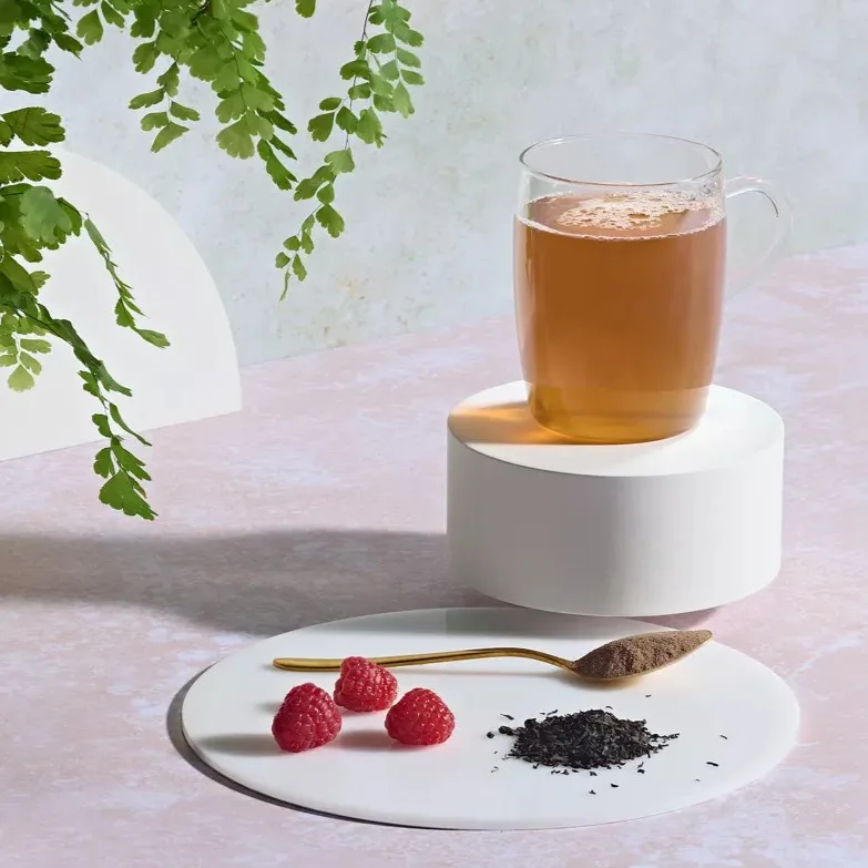 Herbalife instant herbal beverage tea served in a clear glass mug on a white podium pedestal, with fresh raspberries and a gold spoon of tea powder.
