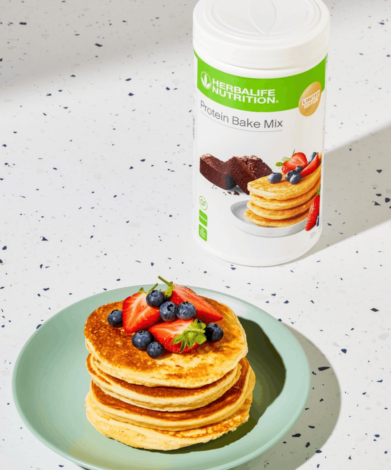 Herbalife Protein Bake Mix tub beside a stack of protein pancakes topped with strawberries and blueberries on a green plate