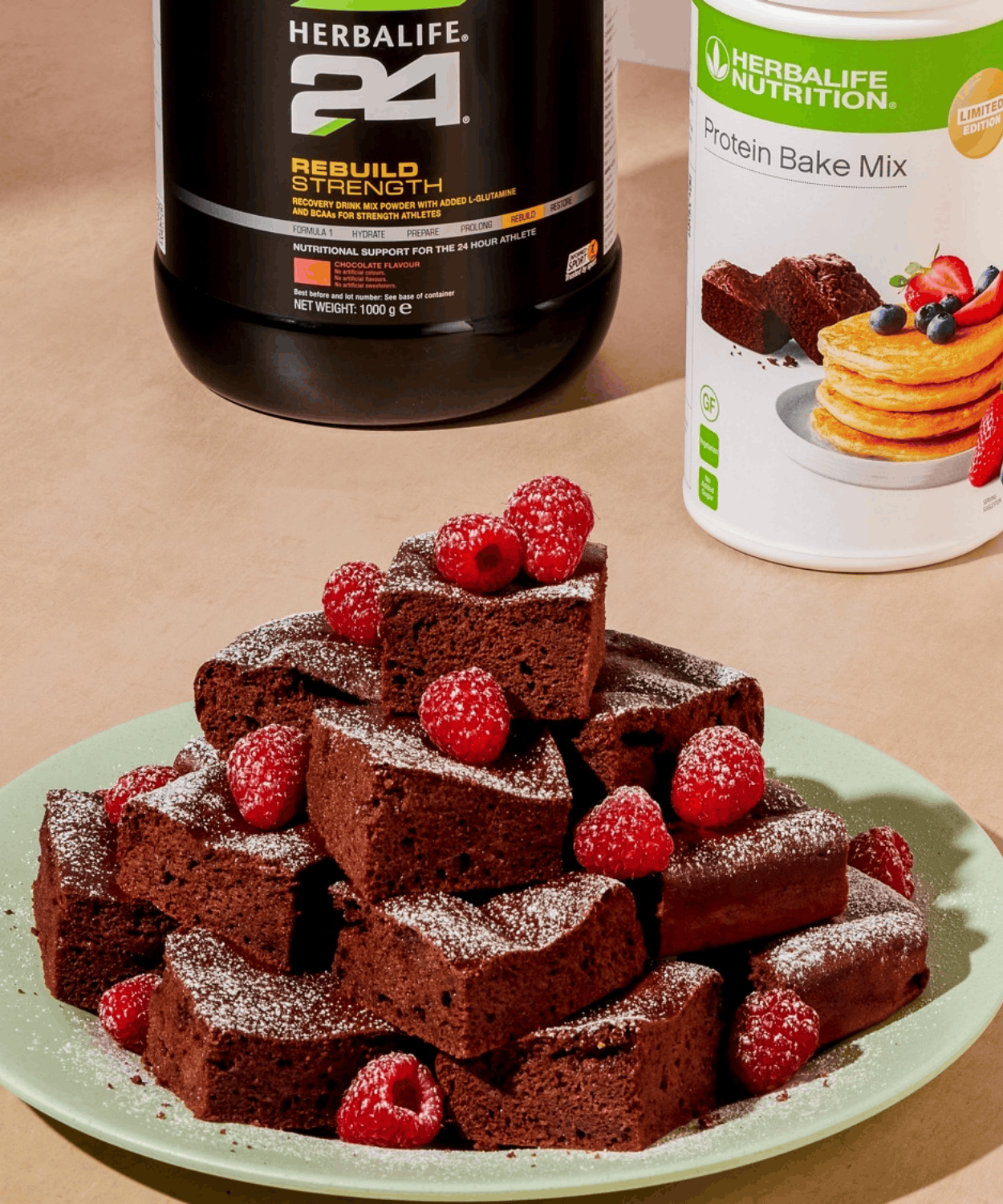 A stack of chocolate protein brownies topped with fresh raspberries, beside Herbalife24 Rebuild Strength and Protein Bake Mix tubs