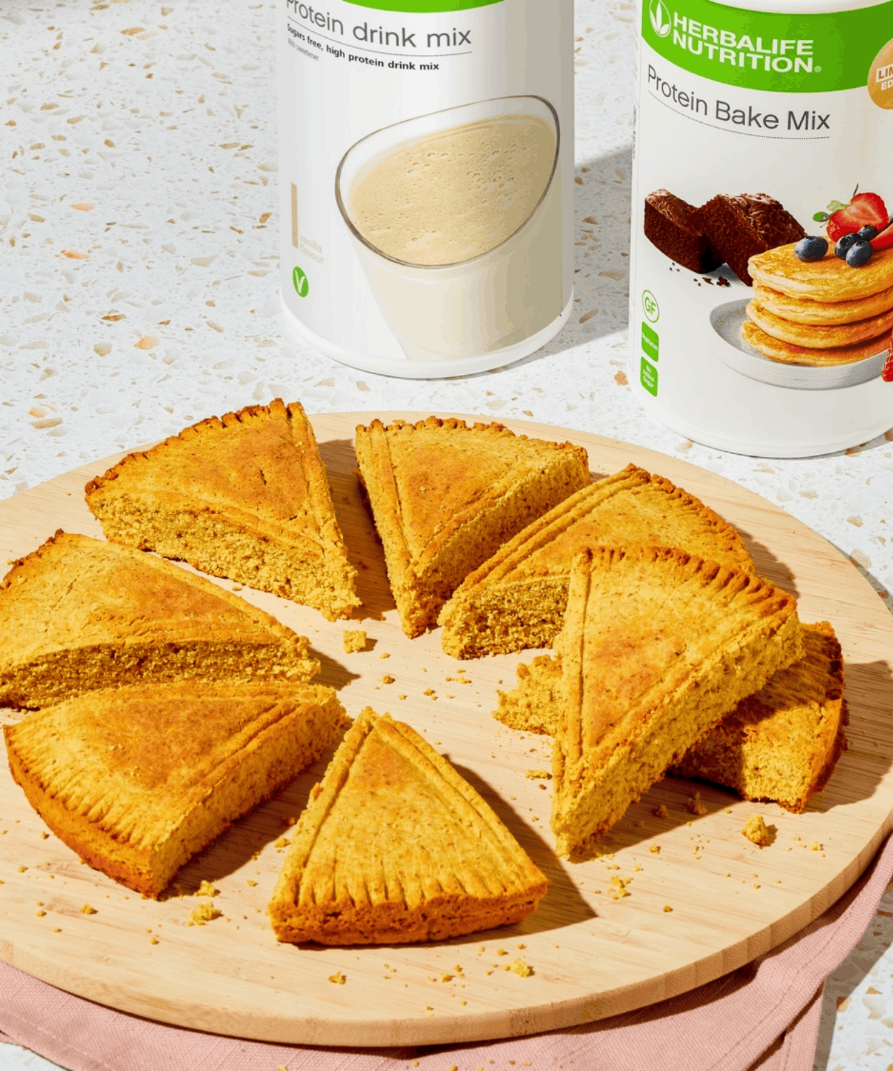 Triangle-cut protein shortbread pieces on a wooden board beside Protein Drink Mix and Protein Bake Mix tubs