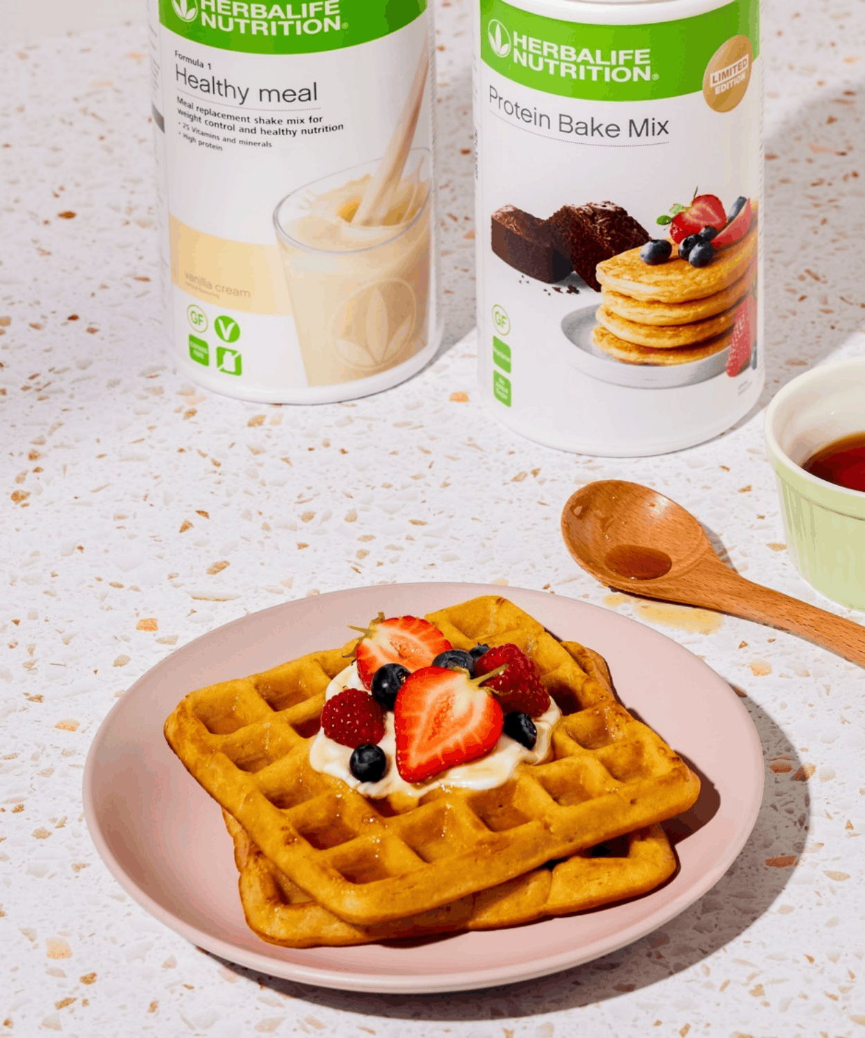 A protein waffle topped with cream, strawberries, blueberries and raspberries on a pink plate, beside Formula 1 Vanilla Cream and Protein Bake Mix tubs