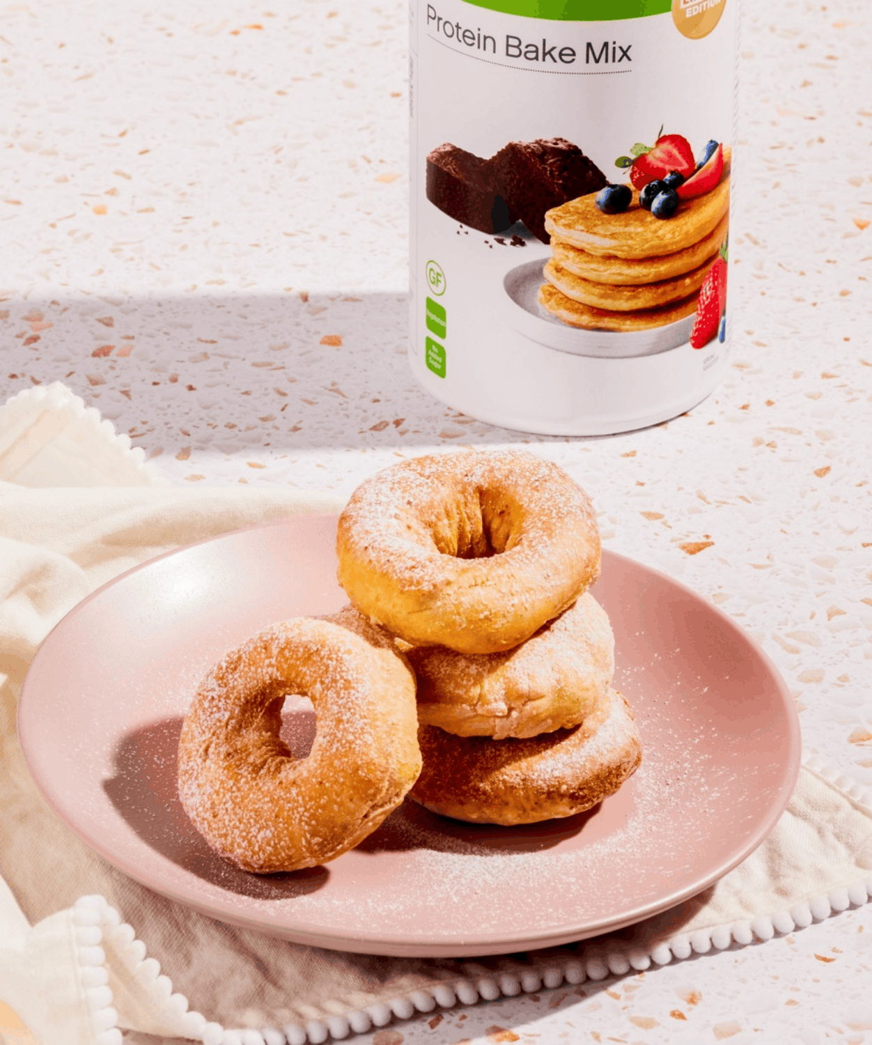 A stack of powdered sugar protein donuts on a pink plate beside the Herbalife Protein Bake Mix tub
