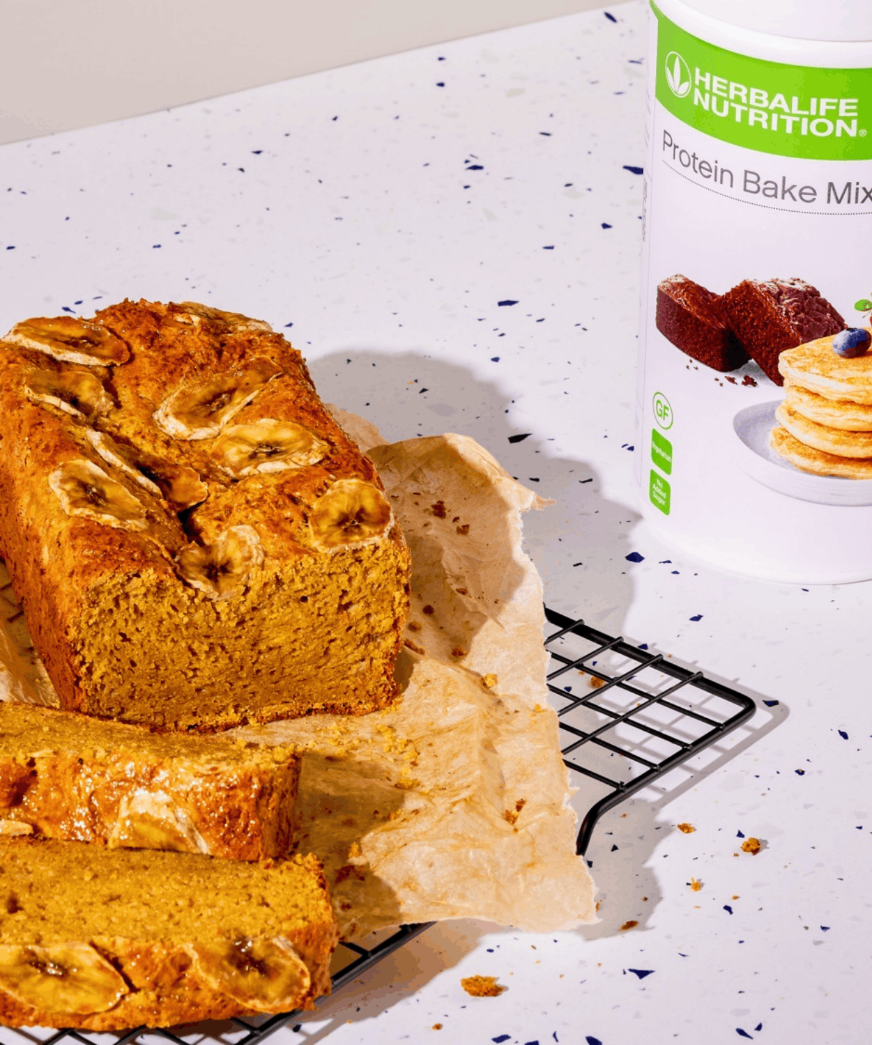 A sliced banana bread loaf topped with banana slices, beside the Herbalife Protein Bake Mix tub