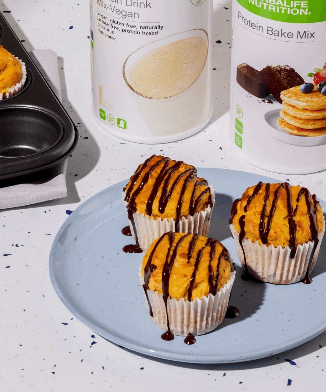 Protein cupcakes drizzled with dark chocolate on a blue plate, beside Protein Drink Mix Vegan and Protein Bake Mix tubs